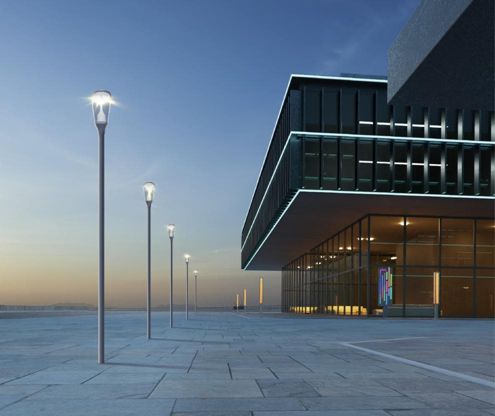 A row of tapered post-top area lighting columns receding along a paved plaza beside a modern glass building at blue hour